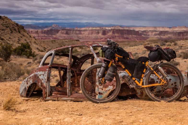 Orange travel bike with black bags in the desert leaning against rusty old body